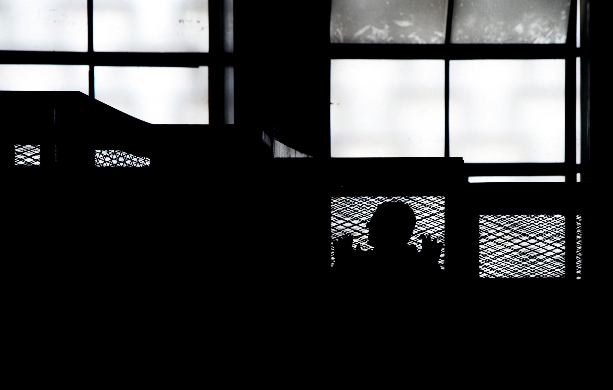 L'immagine presenta una scena in silouette, in cui si intravede la figura di una persona dietro una rete metallica, all'interno di un ambiente che sembra essere un edificio. Le finestre nel fondo lasciano filtrare una luce diffusa, creando un contrasto marcato tra le ombre e la luminosità dello sfondo. L'atmosfera generale risulta suggestiva e misteriosa, enfatizzando la separazione tra la figura e l'ambiente circostante.