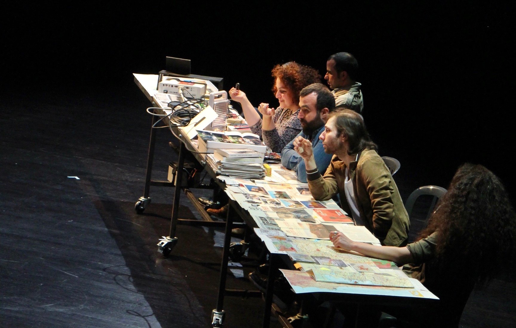 L'image montre un groupe de personnes assises autour d'une grande table sur scène, avec des ordinateurs portables et des documents éparpillés. En arrière-plan, un écran projette un schéma ou une carte, probablement liée à un contexte géographique ou politique, mentionnant des lieux comme Idleb et d'autres noms. Les participants semblent engagés dans une discussion ou une activité collaborative, ce qui suggère un travail de groupe intense. L'ambiance est dynamique, mettant en avant l'importance de l'interaction et de l'échange d'idées.