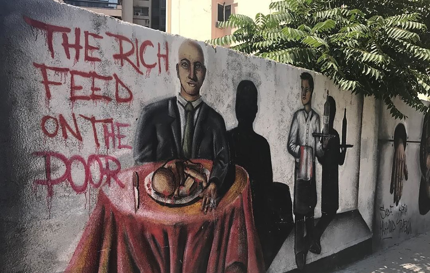 The image features a mural on a wall depicting two main figures. On the left, a man in a suit sits at a table covered with a red tablecloth, holding a loaf of bread and looking directly at the viewer. To the right, another figure dressed as a waiter stands with a tray, while dark shadows cast behind them suggest more figures or a hidden narrative. The phrase "THE RICH FEED ON THE POOR" is painted prominently nearby, conveying a theme of social inequality and critique of class disparity. The background includes urban elements like buildings and greenery, adding to the setting of the mural.