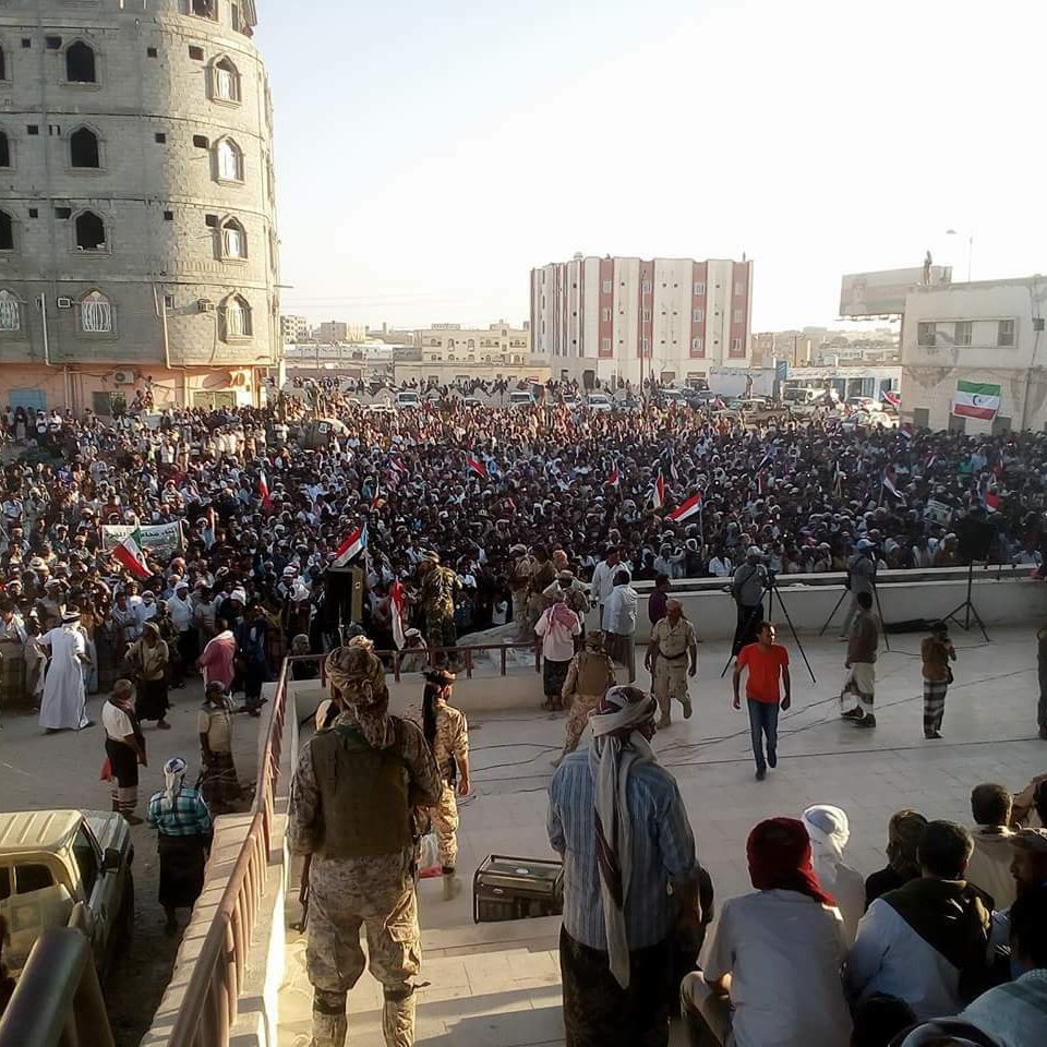 الصورة تظهر تجمعًا حاشدًا من الناس في مكان عام، حيث يبدو أنهم يشاركون في فعالية أو تظاهرة. المجموعة كبيرة وتحتوي على عدد من الأعلام، مما يشير إلى أن هناك موضوعًا معينًا يجمعهم. المباني المحيطة توحي بأن المكان في منطقة حضرية، كما أن بعض الأشخاص يجلسون على الدرج بينما يتواجد آخرون في المقدمة. تبرز الأجواء الحماسية في الحشد.