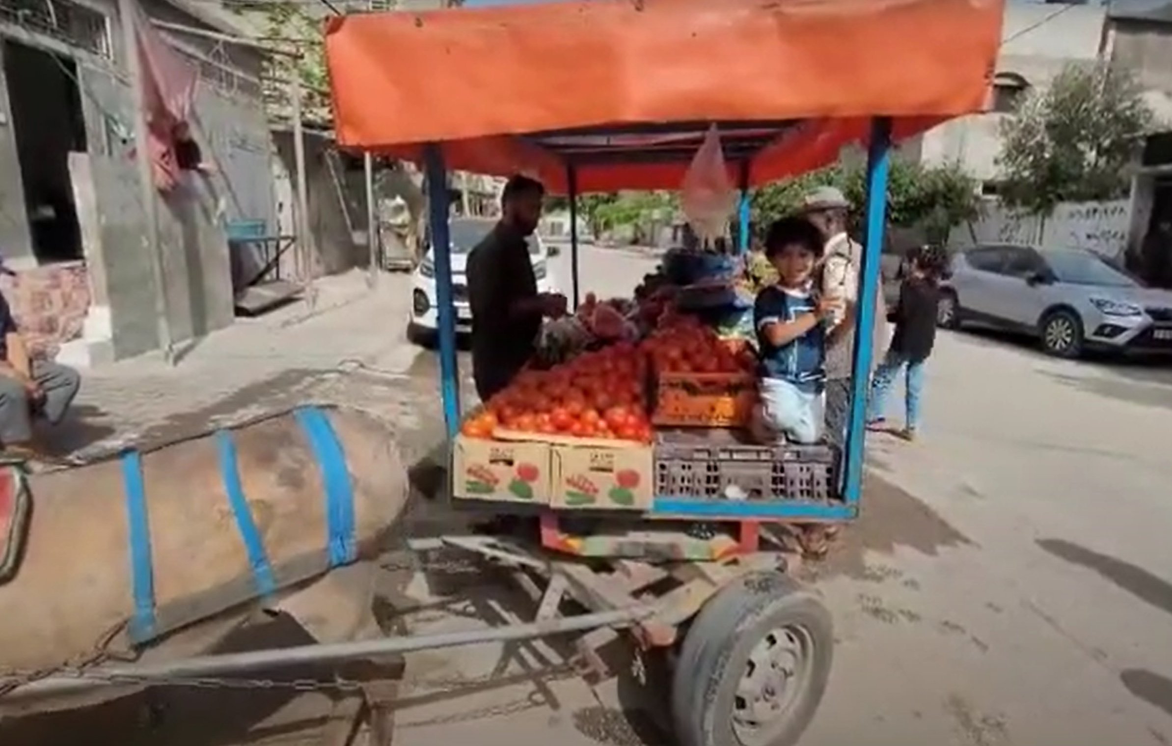 L'image montre un charrette tirée par un âne, pleine de fruits, probablement des tomates. Un homme se tient à côté de la charrette, tandis qu'un enfant est assis sur le bord, tenant des fruits. En arrière-plan, on aperçoit une rue urbaine avec des bâtiments et des personnes. La scène semble animée, typique d'un marché local.