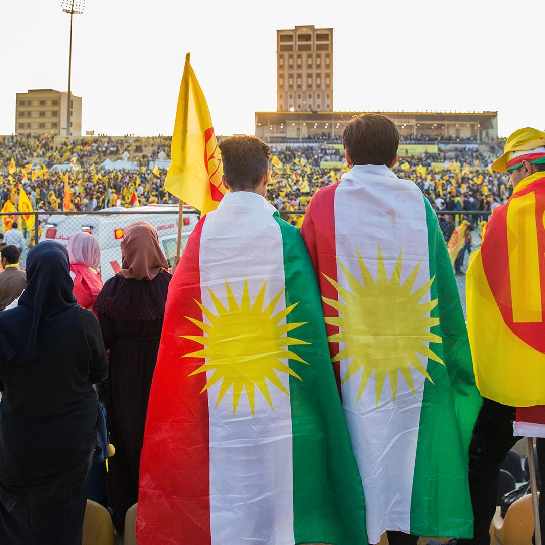 L'image montre un événement festif dans un stade rempli de supporters. Au premier plan, deux personnes portent des drapeaux, l'un étant le drapeau kurde avec les étoiles et l'autre avec un symbole qui semble représenter un mouvement ou un parti. Le public derrière eux est également vêtu de couleurs vives, principalement du jaune, suggérant une ambiance célébratoire. La scène évoque un fort sentiment d'unité et de fierté culturelle.