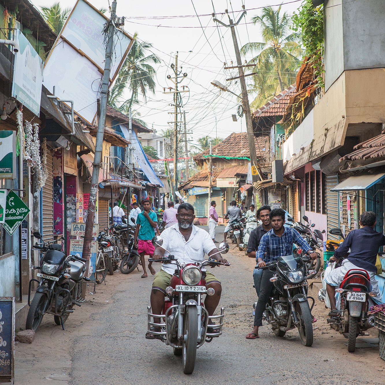 La imagen muestra una calle estrecha en un área urbana, probablemente en un pueblo o ciudad. A ambos lados de la calle hay edificios de un solo piso con techos de tejas. Se pueden ver varios vendedores y tiendas, así como algunas personas caminando y conversando. En el centro de la imagen, un hombre en motocicleta avanza por la calle, mientras que otros motociclistas están estacionados a los lados. La escena refleja la vida cotidiana y el bullicio de un entorno local. También se observa vegetación en el fondo, lo que sugiere un clima cálido y tropical.