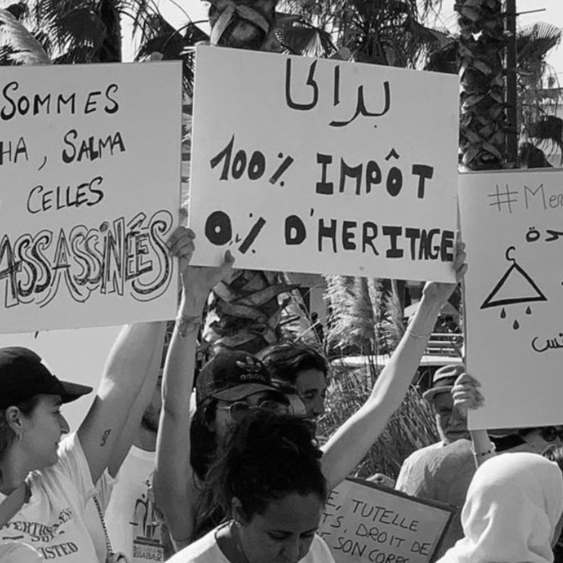 الصورة تظهر مجموعة من الأشخاص يحملون لافتات في مظاهرة. اللافتات تتضمن عبارات تتعلق بالعدالة والحقوق. من بينها نصوص تشير إلى الأحكام الضريبية والمطالبة بالعدالة للضحايا، مما يعكس موقفًا اجتماعيًا أو سياسيًا. الأجواء تبدو حماسية مع حضور العديد من المشاركين.