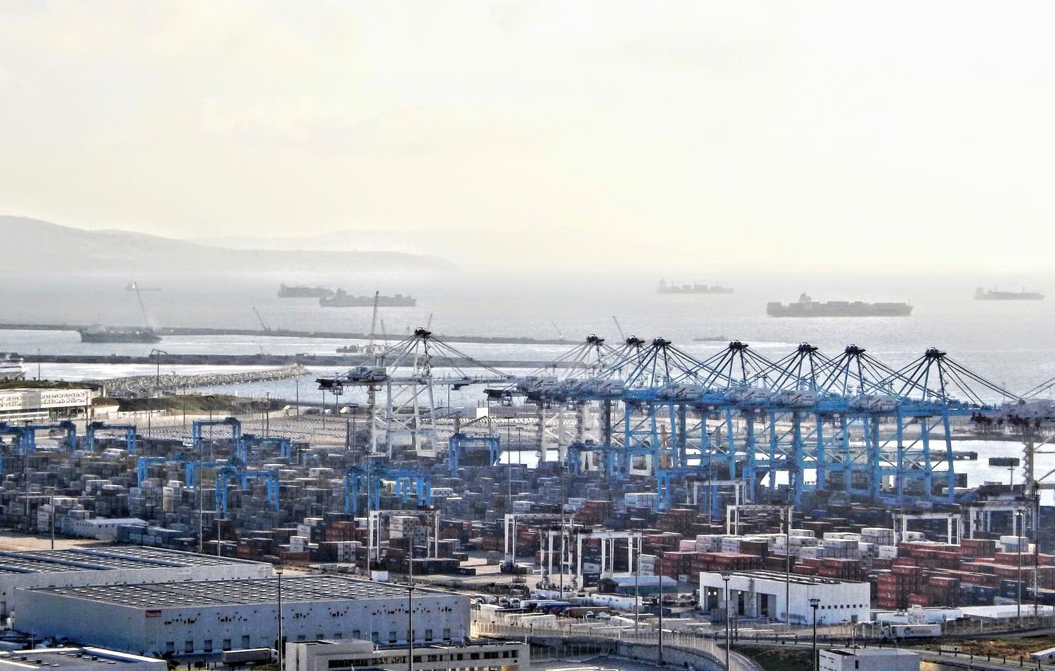 L'image montre un port maritime avec des conteneurs empilés, des grues de chargement et plusieurs navires au large. On peut voir des bâtiments et des installations portuaires. L'horizon est légèrement brumeux, créant une atmosphère calme, et des bateaux de différentes tailles sont visibles sur la mer. L'ensemble dépeint une scène de transport maritime et de logistique.