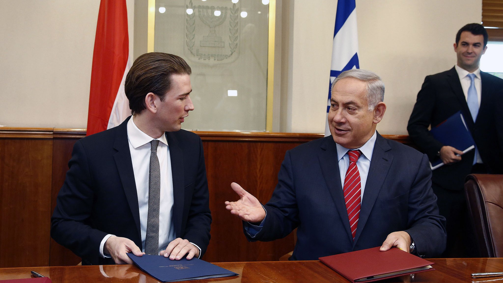 L'image montre deux hommes assis à une table, probablement lors d'une réunion ou d'une cérémonie de signature. L'un d'eux est en costume sombre avec une cravate claire, tandis que l'autre porte une cravate rouge. Ils semblent engagés dans une conversation amicale. À l'arrière-plan, on peut voir des drapeaux, probablement ceux de leurs pays respectifs. L'ambiance générale semble formelle et diplomatique.