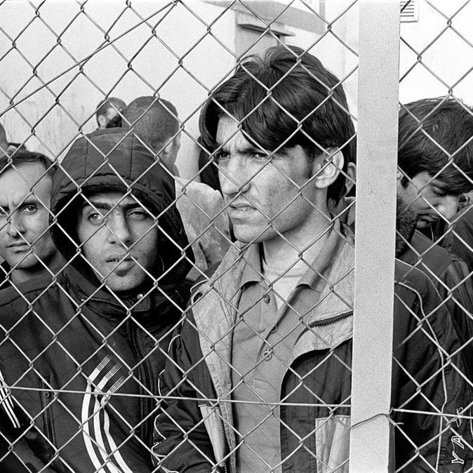 L'image montre un groupe d'hommes, visiblement en attente, qui se trouvent derrière une clôture en fil de fer. Leur expression faciale révèle une certaine anxiété et tension. Ils semblent chercher à voir ou à communiquer avec le monde extérieur. Le cadre en noir et blanc accentue l'intensité de leurs émotions et la gravité de la situation.