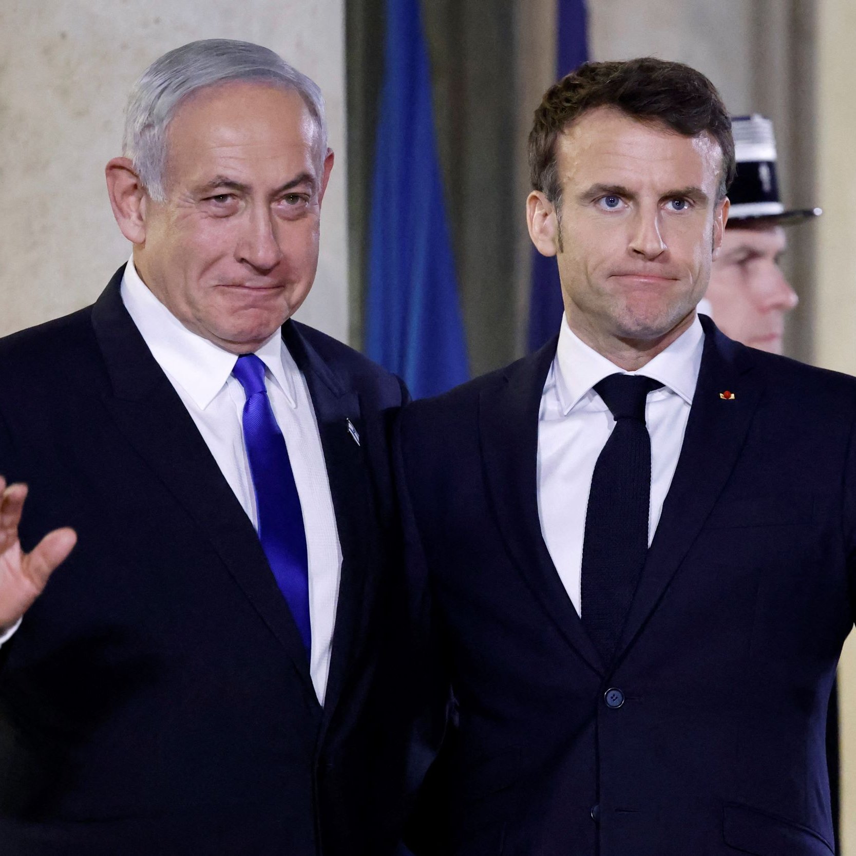 الصورة تظهر شخصين يقفان معًا أمام خلفية مميزة. يبدو أن أحدهما يرتدي بدلة زرقاء وقميص أبيض مع ربطة عنق زرقاء، بينما الآخر يرتدي بدلة سوداء وقميص أبيض مع ربطة عنق داكنة. كلاهما يبتسم قليلاً ويرفعان أيديهما في إشارة ودية. الجو العام في الصورة يبدو رسميًا، مما يشير إلى أن اللقاء قد يكون في مناسبة دبلوماسية أو رسمية.