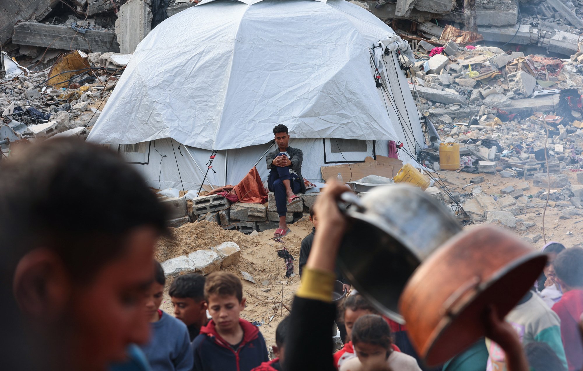 L'image montre un camp de réfugiés après un désastre. Au premier plan, un groupe de personnes autour d'une casserole. En arrière-plan, une tente blanche est visible, avec un jeune assis, regardant son téléphone, tandis que des débris de bâtiments sont éparpillés autour.