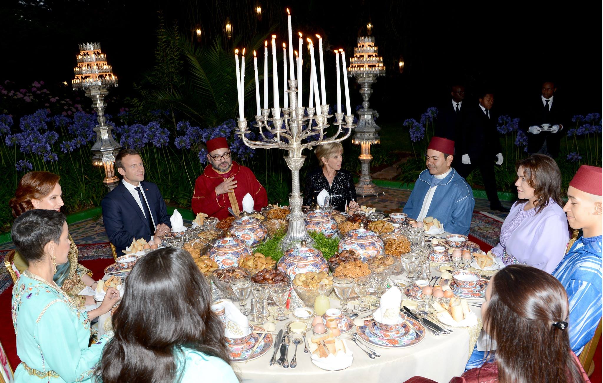 تظهر الصورة مجموعة من الأشخاص حول مائدة رائعة مُعدّة لوجبة طعام فاخرة. المائدة مزينة بأطباق متعددة تحتوي على أطعمة متنوعة، وتحيط بها شمعدانات كبيرة وأجواء أنيقة. يرتدي بعض الضيوف ملابس تقليدية مغربية، بينما يرتدي آخرون ملابس رسمية. تبدو الصورة وكأنها تُعبر عن احتفال خاص أو مناسبة اجتماعية، مع خلفية من النباتات والزهور التي تضفي جوًا جميلًا.