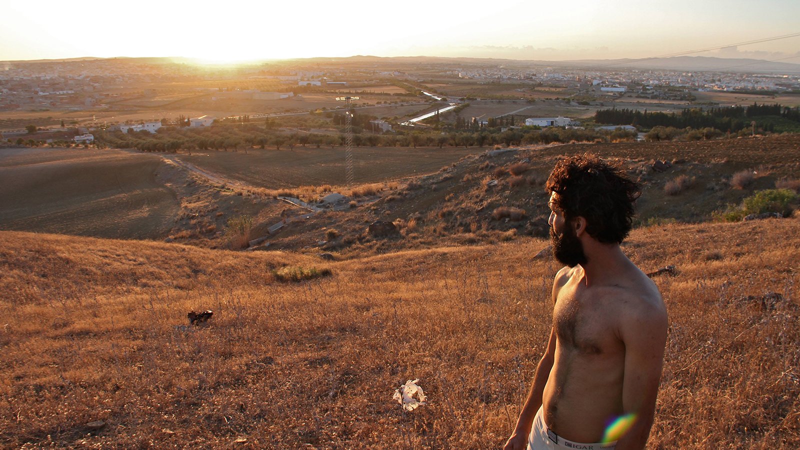 L'image montre un homme debout sur une colline, regardant vers un paysage vallonné au coucher du soleil. Il a les cheveux et la barbe bruns, et porte un t-shirt. Le terrain autour de lui est sec et couvert d'herbes hautes. En arrière-plan, on peut voir un petit village et des champs qui s'étendent à perte de vue, créant un panorama paisible et naturel. La lumière du soleil met en valeur les couleurs dorées de la scène.