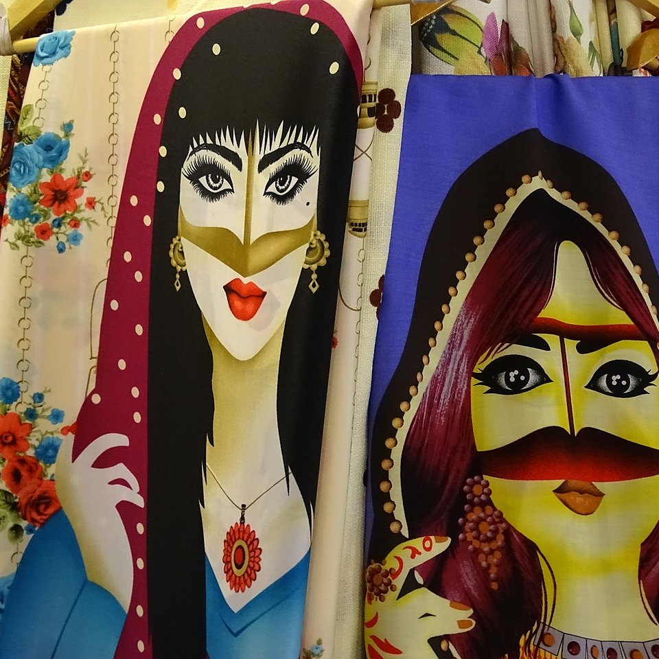 تظهر في الصورة رسومات نسائية ملونة على قماش. الشخصيات المحتواة في الرسومات ترتدي أزياء تقليدية وتتميز بتفاصيل دقيقة في الوجه والعينين. الألوان زاهية، حيث تم استخدام درجات متنوعة من الألوان لجذب الانتباه، مما يعكس ثقافة معينة أو نمط فني مميز. يمكن رؤية بعض الزينة والإكسسوارات حول الشخصيات، مما يضيف لمسة من الأناقة.