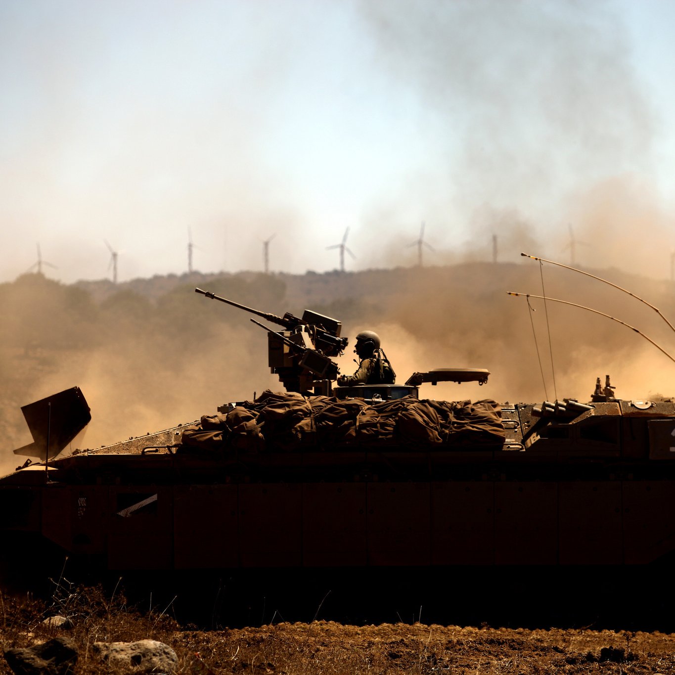 L'image montre un véhicule militaire, probablement un blindé, en action dans un paysage poussiéreux. Un soldat est installé sur le toit du véhicule, armé et prêt. En arrière-plan, on peut apercevoir des éoliennes qui se dressent contre un ciel clair. La scène évoque une ambiance de tension et de préparation militaire, tandis que la poussière soulevée par le mouvement ajoute une dynamique à l'image.