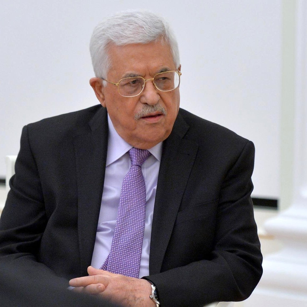 الصورة تظهر رجلًا يجلس في مكان رسمي، يرتدي بدلة سوداء مع قميص أبيض وربطة عنق بنفسجية. لديه شعر أبيض ومصفف بشكل جيد، كما يرتدي نظارات. يبدو أنه في جلسة أو اجتماع حيث تعكس تعابيره الجدية والتركيز. الخلفية بسيطة وفاتحة اللون.