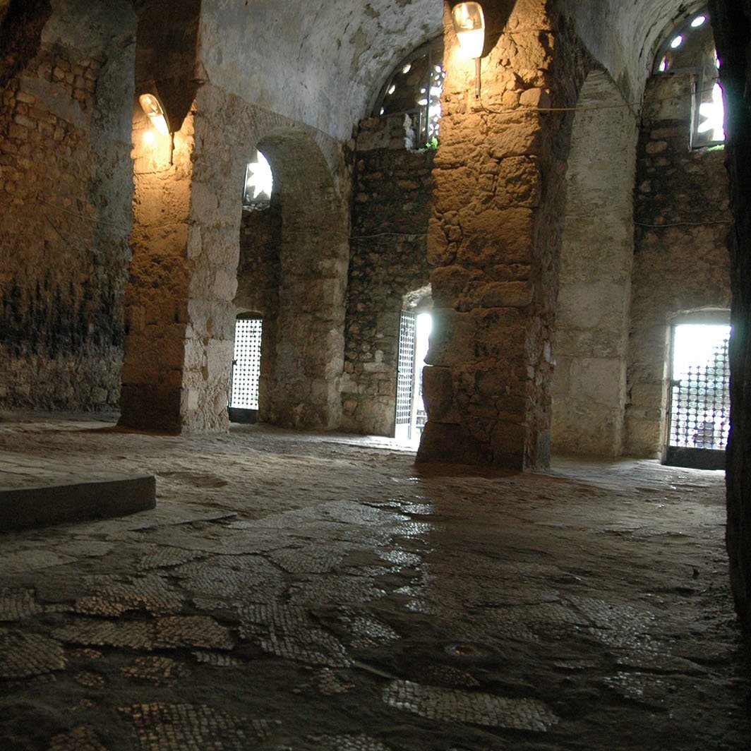 الصورة تُظهر مكانًا داخليًا يرجح أن يكون غرفة أو قبو بُني من الحجر. الجدران مكونة من حجارة كبيرة وغير منتظمة، وتعلوها بعض النوافذ المصفحة بالحديد مما يسمح بدخول الضوء الطبيعي. الأرضية مرصوفة بحجارة صغيرة، وقد تبدو رطبة بعض الشيء. الإضاءة في المكان خافتة، مما يضفي جوًا غامضًا على المكان. يمكن رؤية أجزاء من هيكل قديم يبدو أنه قديم جدًا.