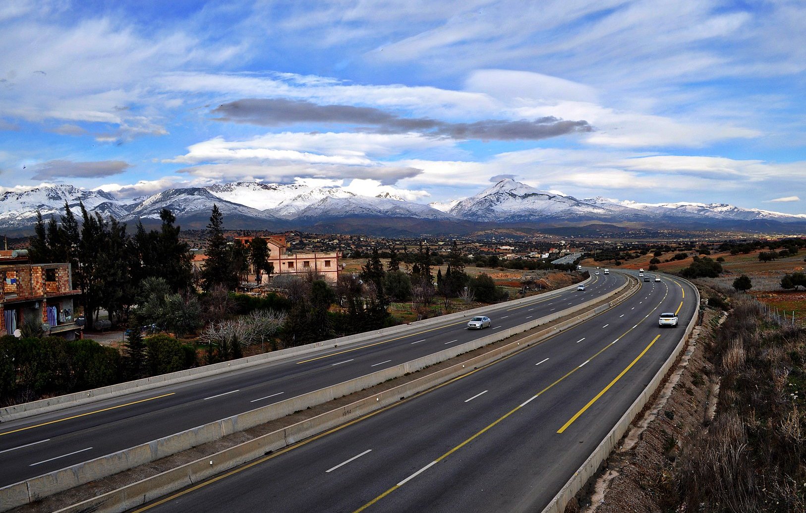 L'image montre une route large et bien entretenue, qui s'étend à travers un paysage pittoresque. On peut voir des montagnes majestueuses au loin, avec des sommets enneigés qui contrastent avec le ciel bleu parsemé de nuages. De chaque côté de la route, il y a quelques bâtiments et de la végétation, ce qui ajoute à la beauté de la scène. Cette vue évoque une ambiance paisible et naturelle, avec un mélange de modernité et de paysages ruraux.