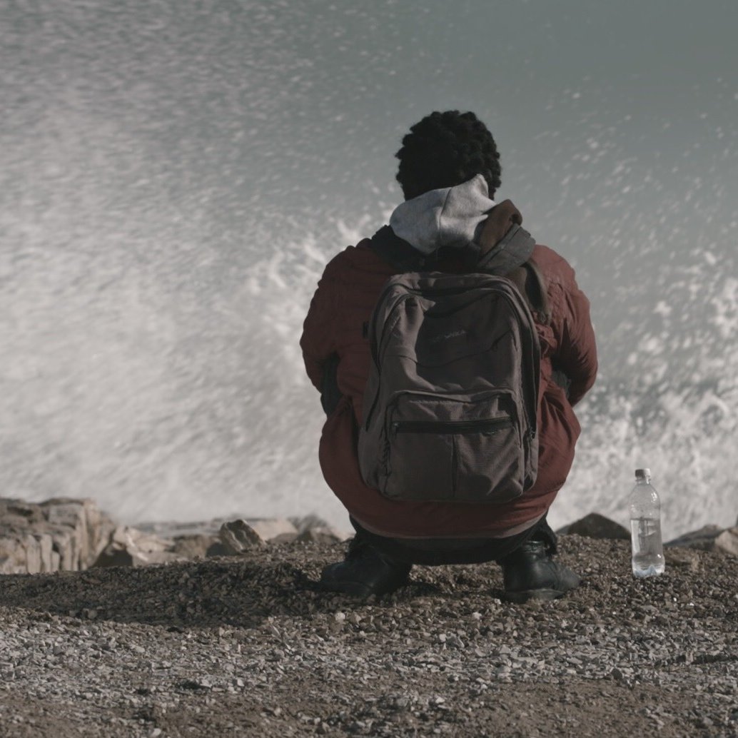 في الصورة، يظهر شخص يجلس على حافة شاطئ أو منطقة قريبة من البحر. يبدو أنه شاب ذو شعر مجعد، ويرتدي معطفًا سميكًا مع كيان داخلي ورأسه مائل قليلاً إلى الأمام. خلفه، تتلاطم أمواج البحر بقوة، مما يعطي إحساسًا بالحركة والطبيعة القوية. بجانبه، يوجد زجاجة ماء على الأرض. الأجواء تبدو هادئة بالرغم من قوة الأمواج، مما يضفي شعورًا بالتأمل أو الوحدة.