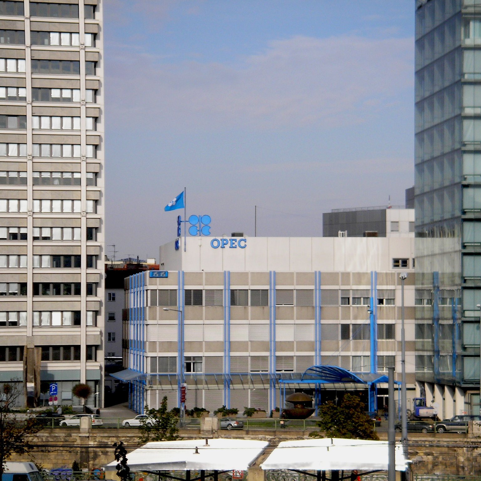 L'image montre le bâtiment de l'OPEP (Organisation des pays exportateurs de pétrole), qui est un bâtiment moderne à plusieurs étages. Des drapeaux sont visibles sur le toit, y compris celui de l'OPEP. Le bâtiment est entouré par d'autres structures urbaines, reflétant un environnement de ville. Le ciel est légèrement nuageux, suggérant une journée calme. Des éléments architecturaux en verre et en béton contrastent avec le design du bâtiment de l'OPEP.