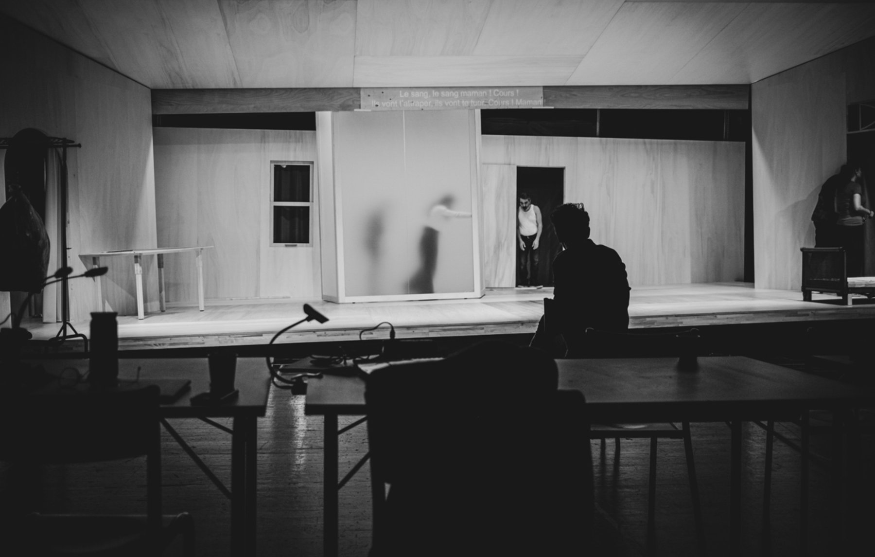 L'image montre un espace scénique avec des murs en bois clair et un éclairage tamisé. On peut apercevoir une silhouette floue à l'arrière-plan, probablement en train de se déplacer. Au premier plan, une personne est assise de dos à une table, observant la scène. L'ambiance est sombre et intimiste, suggérant un moment d'attente ou de concentration avant une représentation. Les éléments de décor, comme les fenêtres et un meuble, ajoutent à l'atmosphère théâtrale.