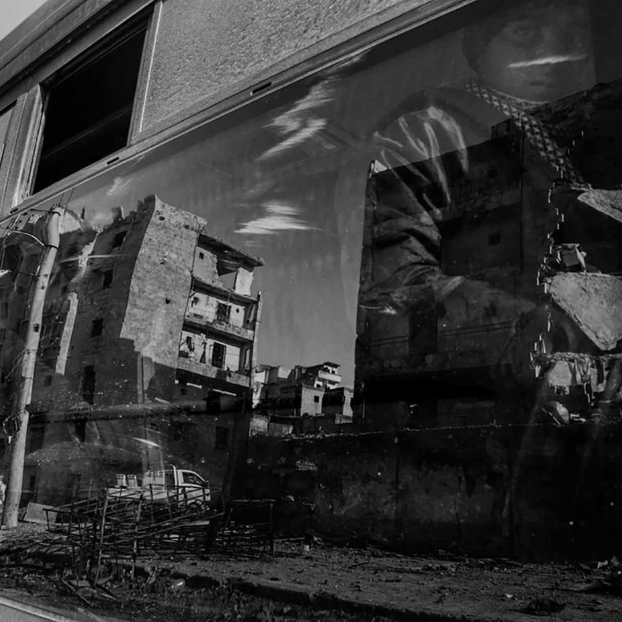 The image features a reflection captured on a glass surface, likely a window or a bus. It shows the distorted but vivid outlines of modern buildings that appear damaged or abandoned, suggesting a scene of urban decay. The reflection also includes a figure partially visible, wearing what looks like a patterned scarf or headdress. The overall tone of the image is monochromatic, emphasizing the somber, evocative nature of the scene. The contrast highlights the interplay between the busy life outside and the desolation depicted in the reflection.