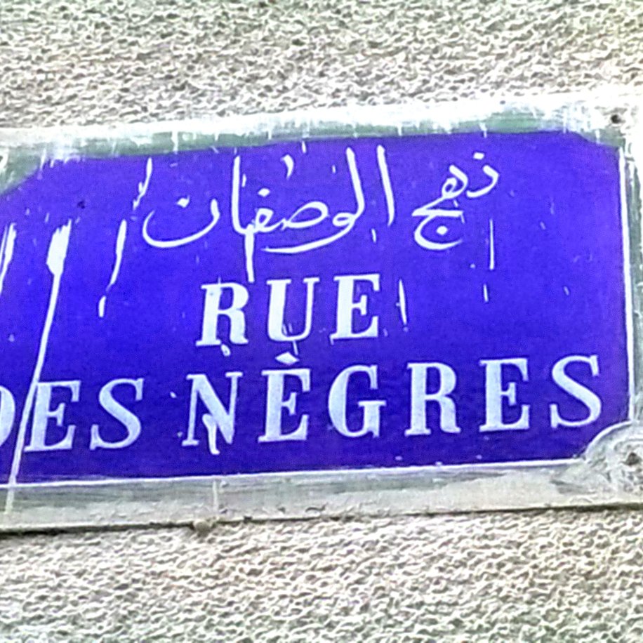 La imagen muestra una placa de calle con un fondo azul oscuro. En la placa se encuentran tres inscripciones en diferentes idiomas: árabe, francés y otro que parece ser un nombre transliterado. La parte central dice "RUE DES NÈGRES" en letras blancas. La placa parece estar un poco desgastada y sucia, lo que indica que ha estado expuesta al tiempo.
