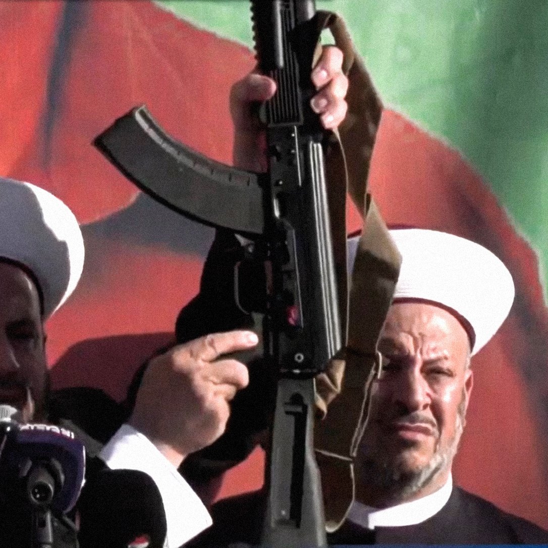 در این تصویر، دو فرد که در حال سخنرانی هستند، به نظر می‌رسد در یک مراسم یا تجمع حضور دارند. یکی از آن‌ها سلاحی را در دست دارد و دیگر فرد در سمت راست نیز در حال نظارت یا همراهی با او است. پس‌زمینه تصویر شامل رنگ‌های زنده و گرافیکی است. هر دو فرد کلاه‌های مخصوص خود را بر سر دارند که نشان‌دهنده هویت مذهبی یا فرهنگی آن‌ها است. احساس کلی تصویر به نظر می‌رسد جدی و تأکیدی بر موضوعی مهم باشد.