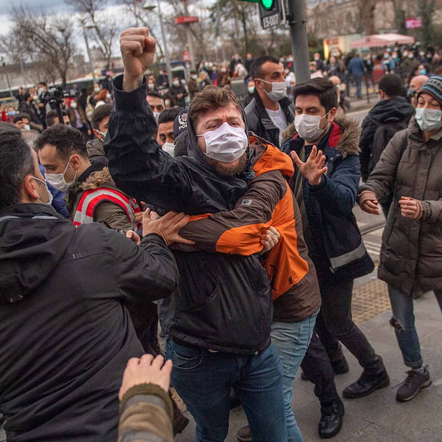 La imagen muestra a un grupo de personas en medio de una situación tensa. Se puede ver a un individuo en el centro, levantando el puño mientras es sujetado por otro, aparentemente en una escena de protesta o manifestación. La mayoría de las personas en la imagen llevan mascarillas. El ambiente parece ser urbano, con un semáforo y algunos edificios de fondo. La expresión de los individuos sugiere emociones intensas, probablemente relacionadas con el conflicto o la agitación social.