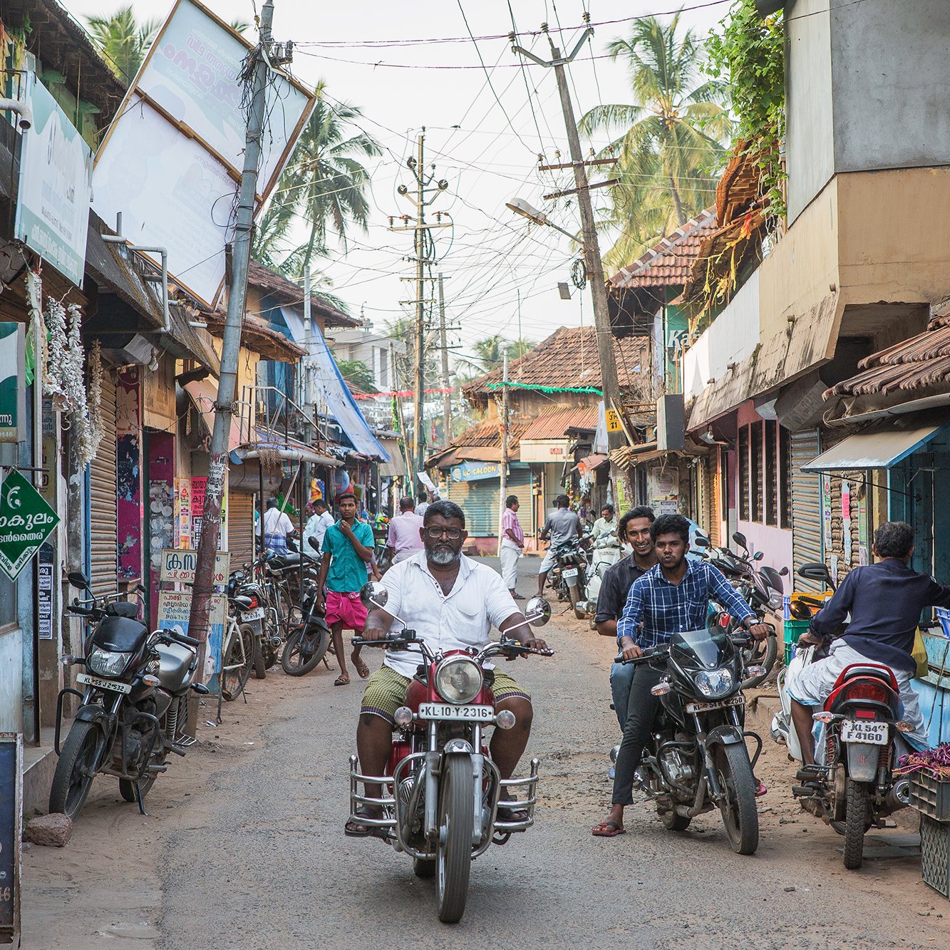 The image depicts a bustling street scene, likely in a South Asian locale. There are narrow, lined pathways with a mixture of residential and commercial buildings, featuring traditional architecture. People can be seen engaging in everyday activities, some on foot and others riding motorcycles. Shops are displaying various goods, and there are signs in a local language. The atmosphere appears lively, with palm trees in the background enhancing the tropical feel of the setting.