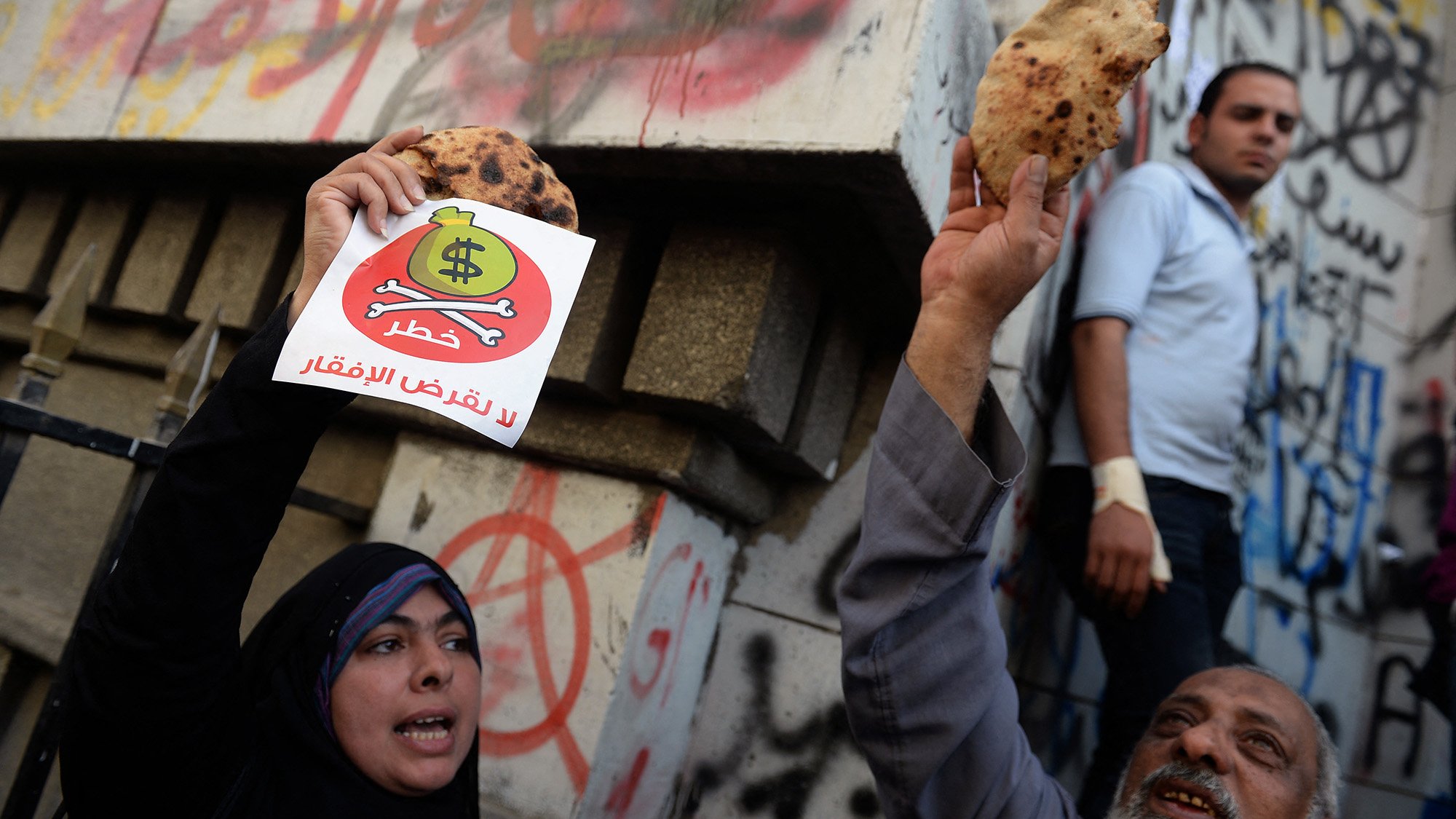 الصورة تظهر مجموعة من الأشخاص في تظاهرة. في المقدمة، يوجد رجل وامرأة يحملان لافتات. المرأة ترتدي الحجاب وتبدي حماسًا بالرفع لافتة مكتوب عليها شعار، بينما الرجل يحمل قطعة خبز. في الخلفية، تظهر جدران مكتوبة عليها شعارات وشعارات فنية. يبدو أن المتظاهرين يعبرون عن رأيهم حول موضوع معين، وملابسهم تشير إلى أجواء الاحتجاج.