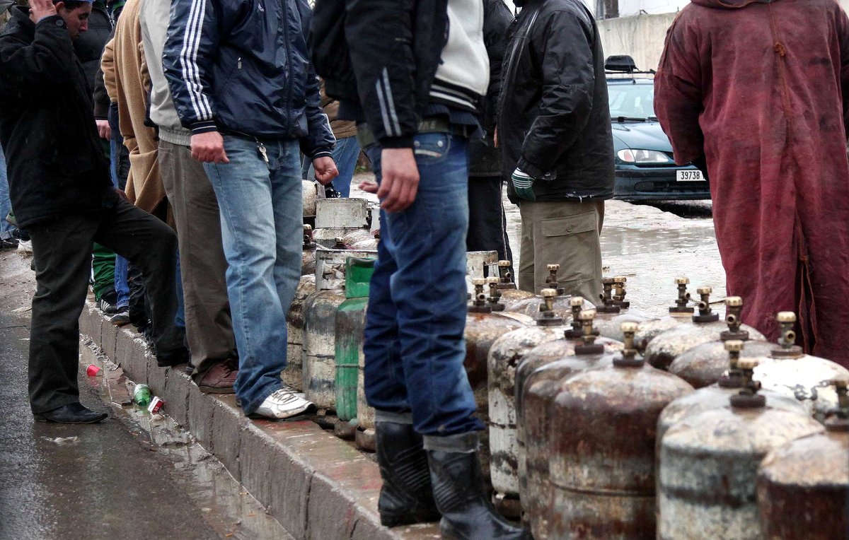 L'image montre un groupe de personnes rassemblées sous la pluie, attendant près de bouteilles de gaz. Certains portent des vêtements imperméables, comme des vestes et des manteaux, tandis que d'autres se protègent de la pluie avec des parapluies. L'atmosphère semble sombre et humide, suggérant une situation de manque ou d'attente. En arrière-plan, des voitures sont visibles, ainsi que des bâtiments. Les gens semblent engagés dans une activité collective, probablement en attendant d'acheter du gaz.