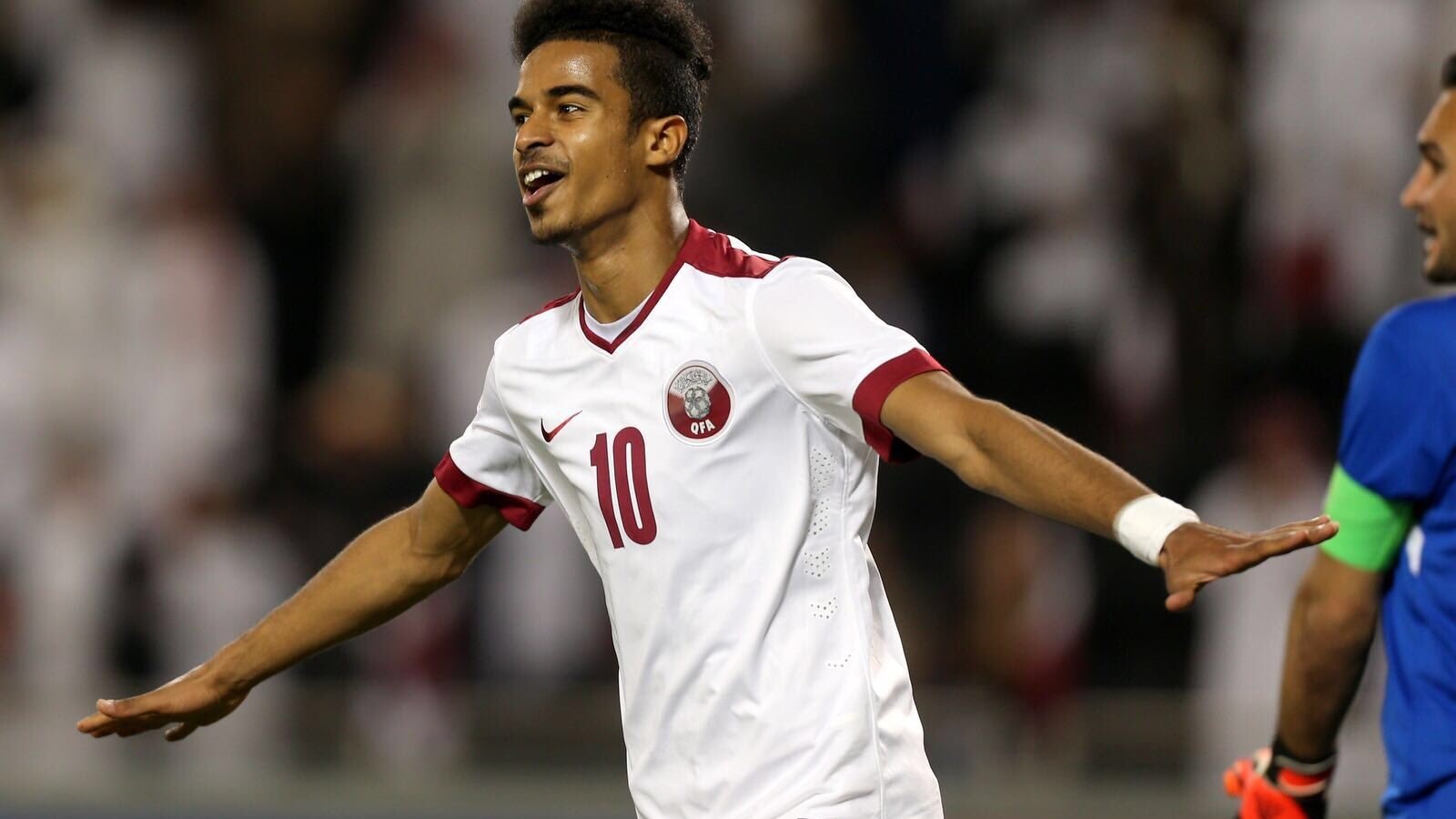 L'image montre un jeune joueur de football qui célèbre un but. Il porte un maillot blanc avec des touches de rouge, et le numéro 10 est visible sur son dos. Son expression est joyeuse et dynamique, manifestant sa satisfaction après une action réussie. En arrière-plan, on aperçoit des supporters, probablement vêtus de blanc, ce qui suggère un événement sportif important. La scène dégage une énergie positive et une intensité propre au football.