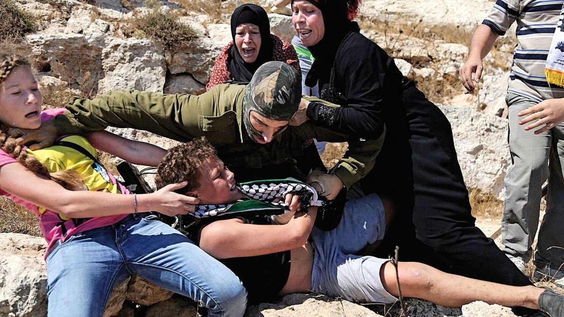 L'image montre une scène de tension où plusieurs personnes semblent être impliquées dans une confrontation. Un militaire en uniforme est en train de maîtriser un jeune homme au sol tandis que d'autres personnes, dont des femmes et des enfants, semblent réagir avec de l'indignation ou de la peur. L'environnement paraît rural, avec des rochers et une végétation sèche. L'émotion et le conflit sont palpables dans cette image.