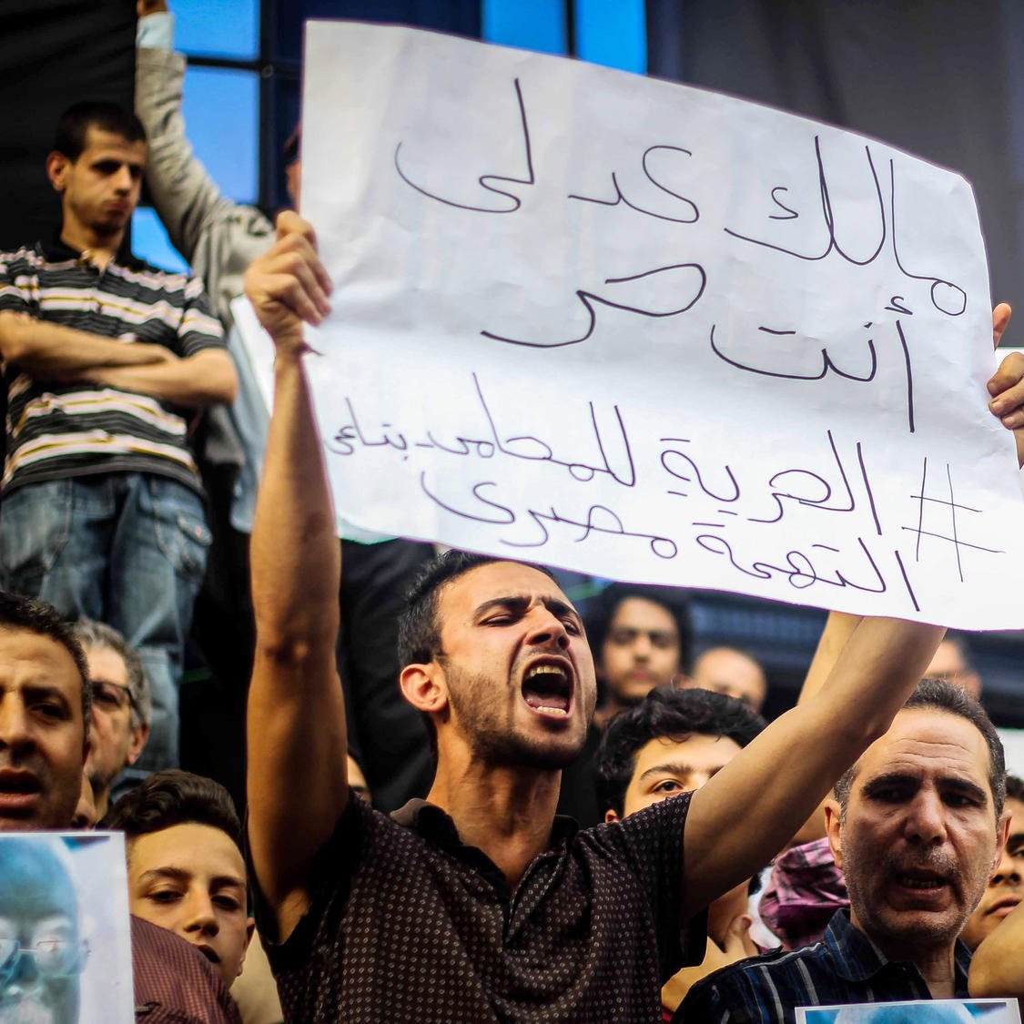 The image depicts a crowd of protestors holding signs during a demonstration. One individual is prominently featured, passionately expressing their views. The sign reads a message in Arabic, which translates to a call for freedom and justice. The crowd appears engaged and determined, highlighting the intense atmosphere of the protest. Various faces in the background show a mix of emotions, reflecting the seriousness of the situation.
