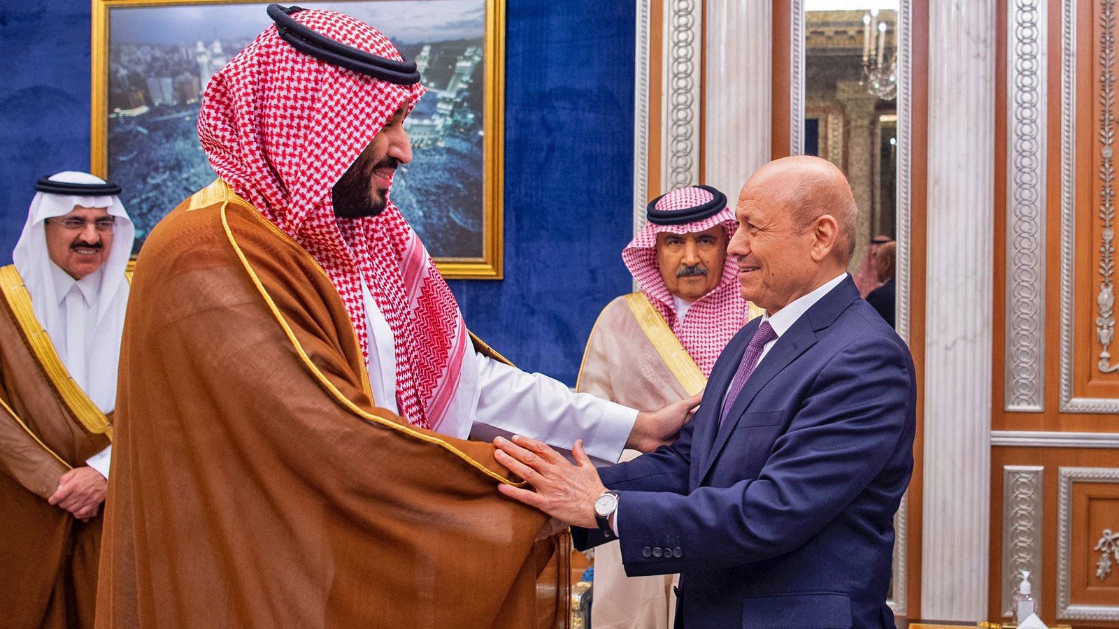 L'image montre deux hommes en costume, engagés dans une interaction amicale, probablement lors d'une rencontre officielle. L'un des hommes porte un habit traditionnel avec un châle. L'arrière-plan présente un décor élégant, typique d'un palais, avec des éléments architecturaux raffinés. D'autres personnes en tenue formelle sont visibles, suggérant un contexte diplomatique ou cérémoniel.