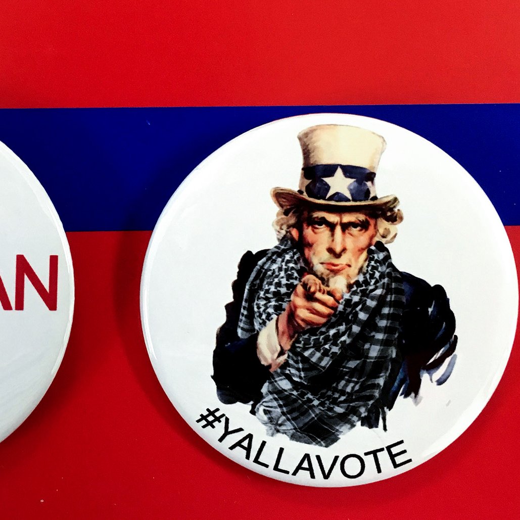 L'image montre deux badges sur un fond rouge avec une bande bleue. Le premier badge mentionne "ARAB AMERICAN VOTER" avec un symbole de coche, tandis que le deuxième badge représente un personnage qui ressemble à l'oncle Sam, avec le texte "#YALLAVOTE". Ces badges mettent en avant l'importance du vote et de l'engagement civique, en particulier au sein de la communauté arabe-américaine.