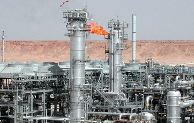 L'image montre une installation industrielle, probablement liée à l'industrie pétrolière ou gazière. On peut voir plusieurs tours métalliques, dont certaines émettent des flammes, indiquant une combustion, peut-être de gaz résiduel. L'arrière-plan présente un paysage désertique, caractérisé par des collines ou des montagnes sèches, typique des zones où se trouvent des installations énergétiques. Les structures sont en métal et semblent sophistiquées, témoignant d'une technologie avancée utilisée dans cette industrie.