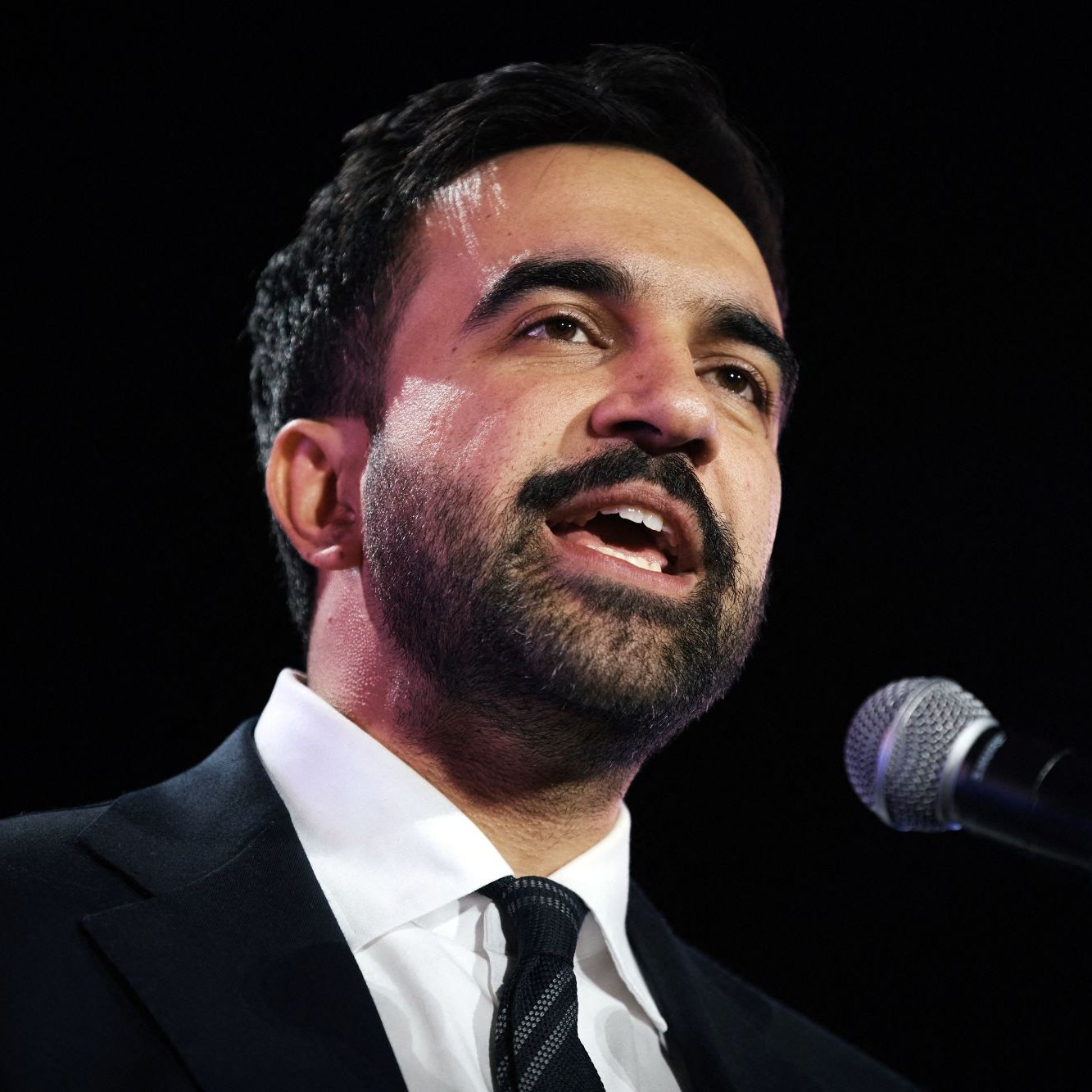 مردی با لباس رسمی، در حال سخنرانی و صحبت در میکروفون است. احساس جدیت و اعتماد به نفس دارد.