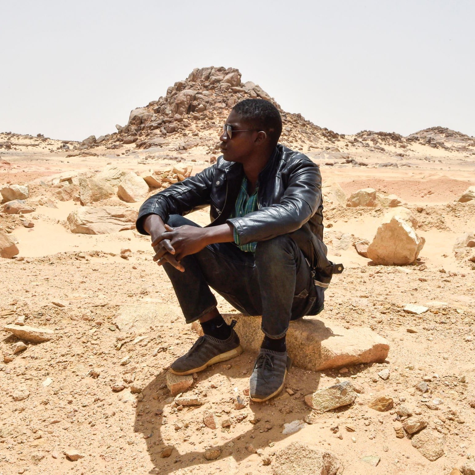 L'image montre deux personnes assises dans un paysage désertique. L'un des hommes est en train de s'asseoir sur une grosse pierre, portant une veste en cuir et des lunettes de soleil. Il semble pensif, observant le paysage autour de lui. En arrière-plan, une autre personne est également assise, mais davantage éloignée. Le terrain est constitué de sable et de roches, avec des formations rocheuses au loin, sous un ciel clair. L'atmosphère semble calme et isolée.