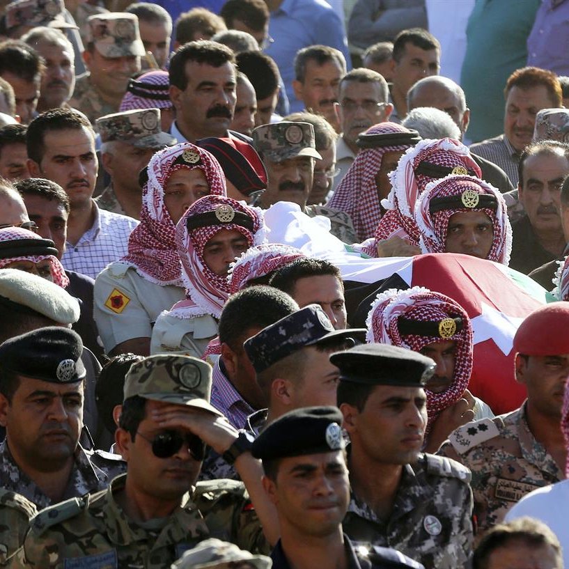 The image depicts a large group of people gathered in a public setting for a somber occasion. In the foreground, several individuals are wearing traditional attire, including headscarves and distinctive clothing, which suggests a cultural or regional significance. They appear to be carrying a casket draped in a flag, indicating a funeral or memorial service. The crowd encompasses military personnel dressed in uniforms, highlighting a sense of respect and ceremony. The atmosphere is likely one of mourning and solidarity as attendees pay their respects.