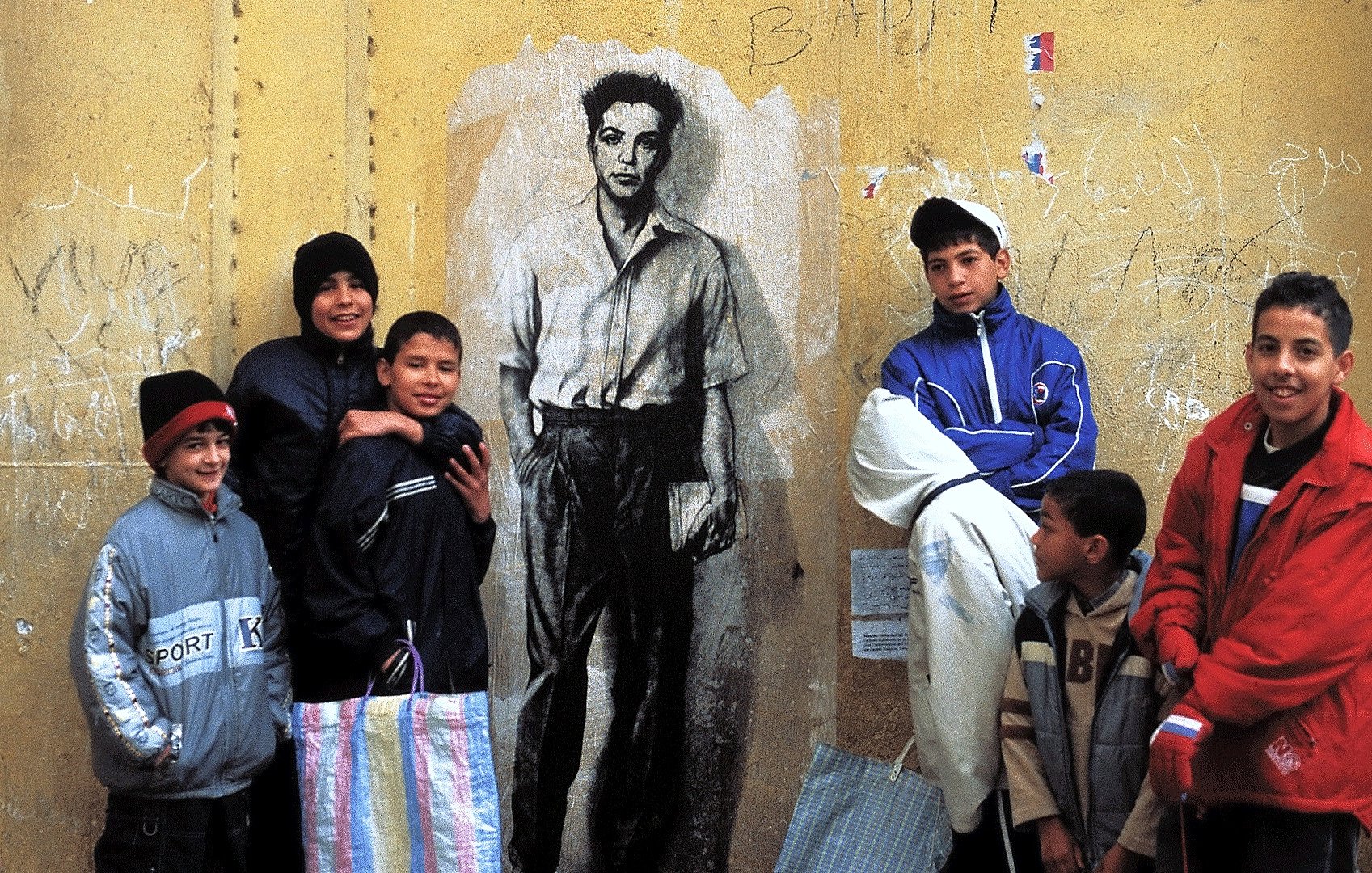 The image features a group of children standing in front of a large mural on a yellow wall. The mural depicts a black-and-white portrait of a young man in a shirt. The children are dressed in casual, colorful clothing, with some wearing hats. They appear to be interacting playfully, with a sense of camaraderie among them. The background shows some graffiti and markings, adding to the urban setting.