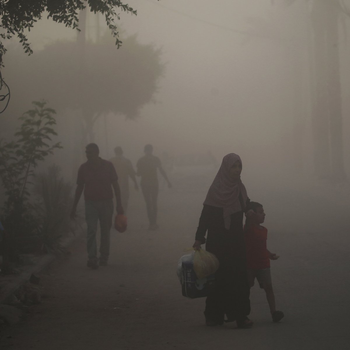 L'immagine mostra una scena di una strada avvolta nella nebbia o nella polvere. In primo piano, una donna con un velo scuro tiene la mano di un bambino, mentre camminano insieme. Sullo sfondo, si possono vedere altre persone che camminano, ma sono poco visibili a causa della foschia. Gli alberi e alcune piante ai lati della strada creano un'atmosfera un po' opprimente. La luce sembra attenuata, dando un'impressione di desolazione.