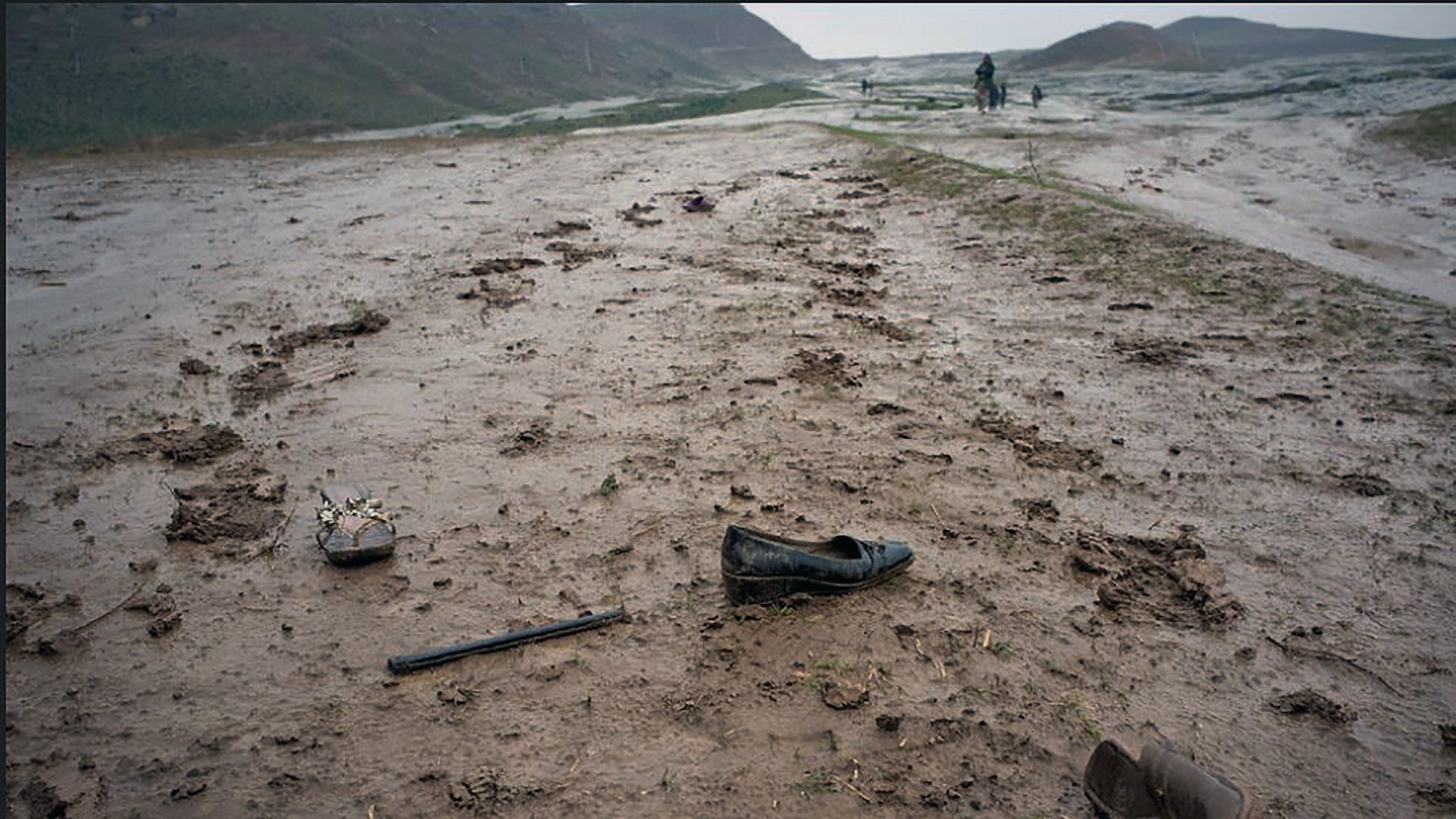 The image depicts a muddy landscape, likely after rainfall. The ground is uneven and dotted with puddles and dirt. Scattered across the terrain are several shoes, including a black dress shoe and what appears to be a wooden shoe or clog. There is also a long, thin object that resembles a cane or stick. The scene conveys a sense of abandonment and desolation, with lush green hills in the background under a gray sky.
