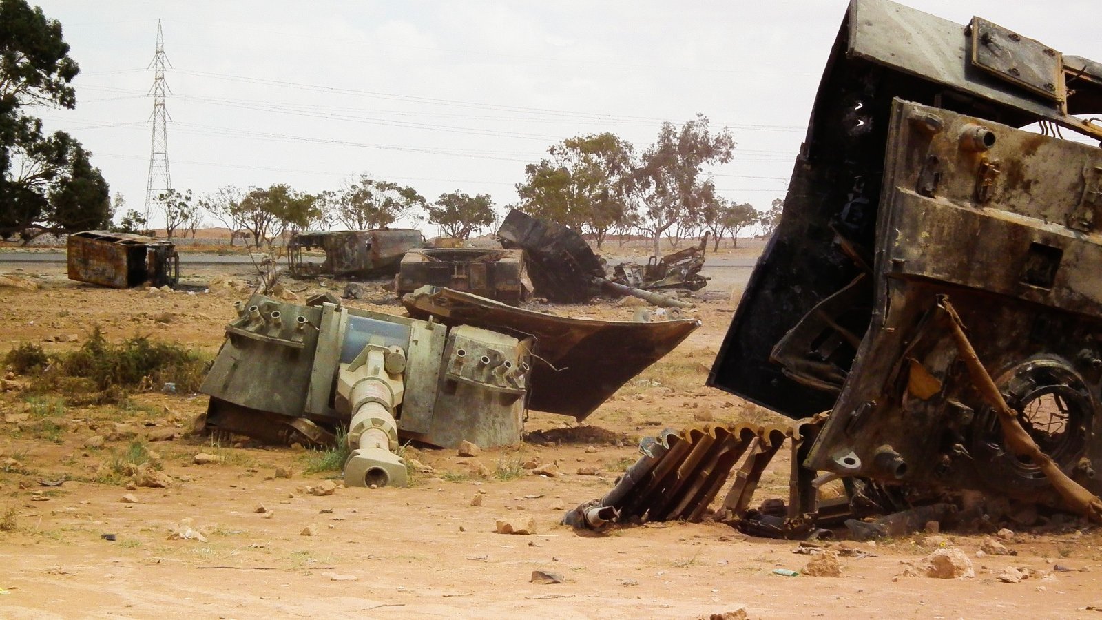 L'image montre un paysage désertique où sont dispersés des véhicules militaires rouillés et abandonnés. On peut voir des carcasses de tanks et d'autres machines, gisant sur le sol poussiéreux. Au loin, des arbres clairsemés et des pylônes électriques se distinguent, ajoutant à l'ambiance aride et désolée du lieu. Le ciel est nuageux, ce qui donne une atmosphère grise à la scène.