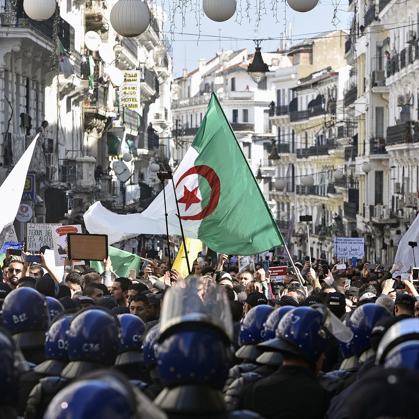 En la imagen se puede ver una multitud reunida en una calle, con varias personas sosteniendo banderas argelinas y carteles. El ambiente parece ser de una manifestación o protesta, ya que hay un grupo de policías en primer plano, equipados con cascos y protecciones, observando la situación. Los edificios a ambos lados de la calle son visibles, creando un contexto urbano y de actividad social.