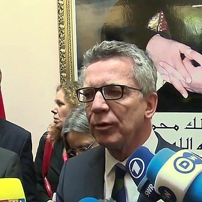 L'image montre un groupe de personnes lors d'une conférence de presse ou d'un événement officiel. Au premier plan, un homme s'exprime devant des caméras et des micros, entouré de plusieurs autres individus qui semblent attentifs. En arrière-plan, on peut voir un portrait ou une image représentant des mains avec des symboles significatifs, ainsi qu'un drapeau. L'atmosphère semble formelle, probablement liée à des discussions d'ordre politique ou diplomatique.