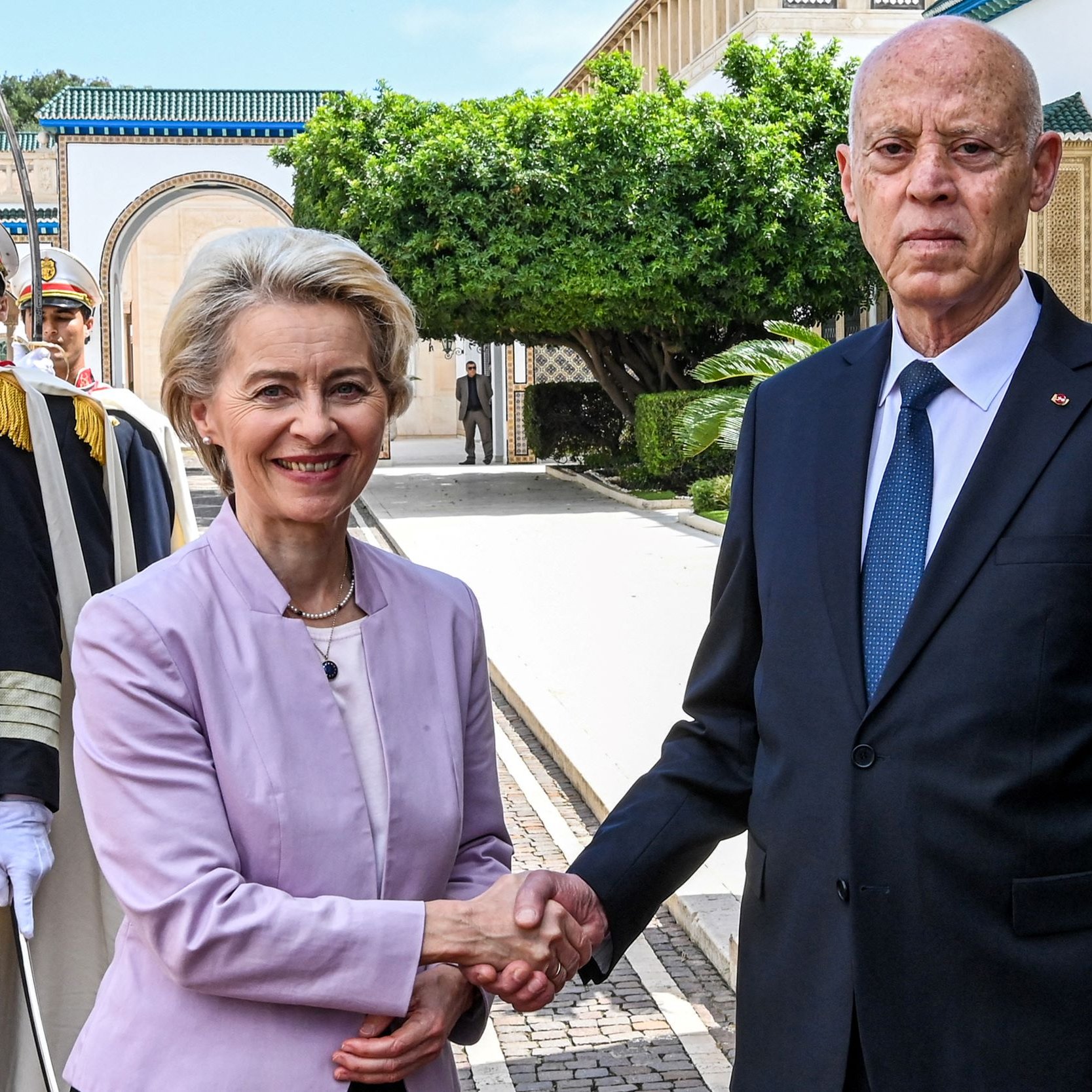 L'image montre une scène formelle où deux personnes se serrent la main. À gauche, une femme souriante porte un costume clair, tandis qu'à droite, un homme en costume sombre semble sérieux. En arrière-plan, on peut voir des gardes en uniforme et des éléments architecturaux d'un bâtiment. La végétation luxuriante ajoute une touche de couleur à la scène. L'atmosphère semble être celle d'une rencontre officielle.