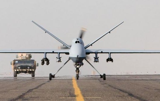 L'image montre un drone militarisé en position de décollage, vu de face, sur une piste d'atterrissage. En arrière-plan, on aperçoit un véhicule militaire qui s'approche. Le ciel semble légèrement nuageux ou brumeux, créant une ambiance de calme avant l'action. Le drone est de couleur claire, avec des ailes larges et un design aérodynamique, prêt à s'envoler.