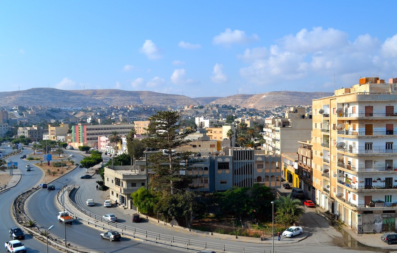 L'image montre un paysage urbain avec des bâtiments variés, y compris des immeubles résidentiels et commerciaux, ainsi qu'une route serpentine. Au loin, on peut voir des collines et un ciel clair avec quelques nuages. La scène dégage une ambiance paisible, avec des voitures circulant sur la route et des arbres ajoutant une touche de verdure à l'environnement.