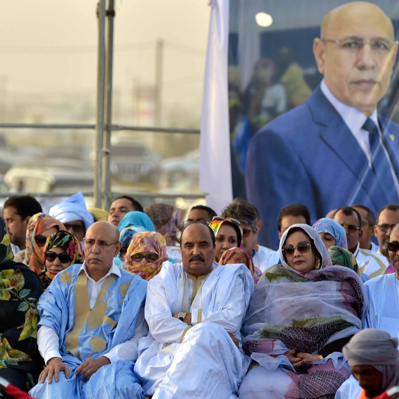 L'image montre un groupe de personnes assises, participant à un événement ou à une cérémonie. Elles portent des vêtements traditionnels, souvent des robes colorées et des turbans. En arrière-plan, il y a un grand portrait d'un homme en costume, probablement une figure politique, qui semble surveiller l'événement. L'ambiance paraît solennelle et impliquée, avec beaucoup d'attention portée au discours ou à la célébration en cours.