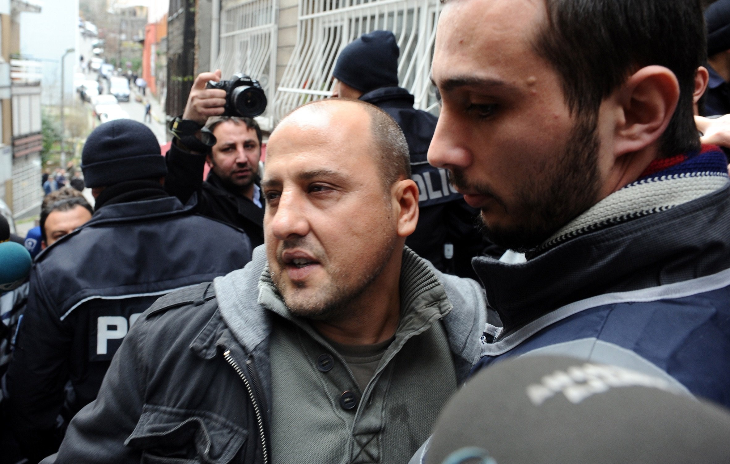 The image depicts a scene with a group of people, likely in a city setting. In the foreground, two men are present. One man, appearing distressed or concerned, is being escorted by another, who seems to be in a position of authority. The background includes police officers and a crowd, indicating a tense situation or an event such as a protest or arrest. The overall atmosphere appears to be one of heightened emotion or urgency.